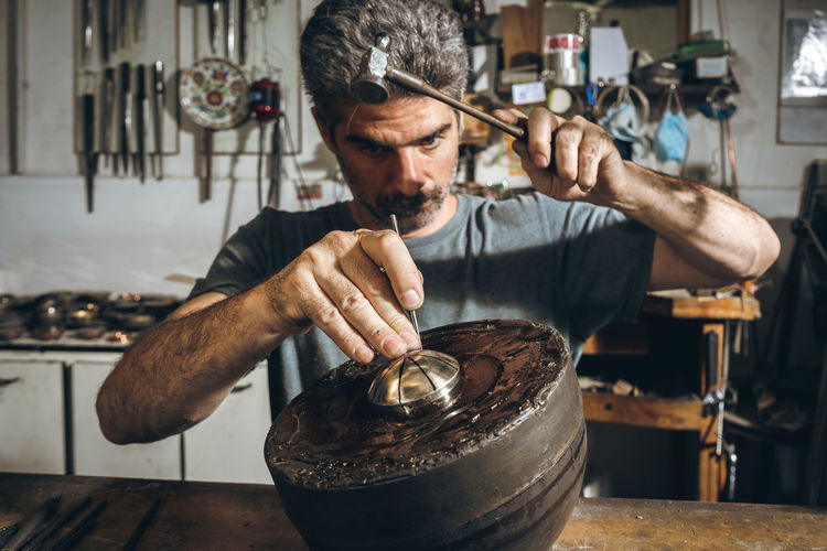 Serious mature male artisan using metal punch and hammer to carve ornament on silver lid while sitting at table in professional workshop