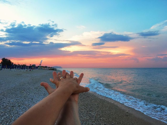 Cropped hands of person by sea against sky during sunset