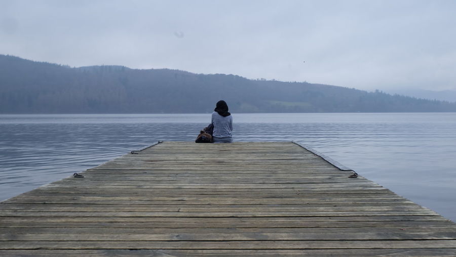 Rear view of woman sitting on pier over lake during foggy weather