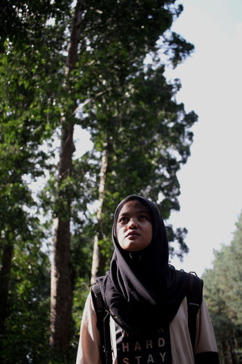Low angle view of young woman in hijab standing against trees