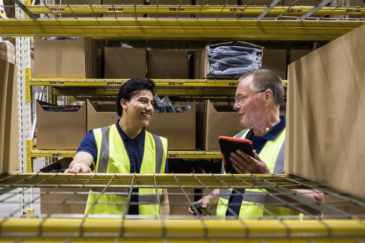 Smiling male coworkers discussing while seen through rack at warehouse