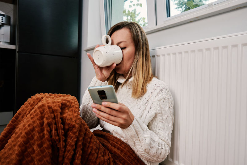 Worried woman sit near heating radiator under blanket and using smartphone