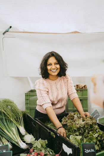 Portrait of smiling female owner standing near vegetable crate in stall at market