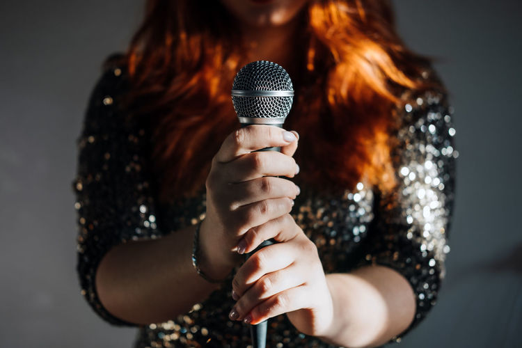 Faceless portrait of redhead woman in sparkly evening dress holding microphone on dark night