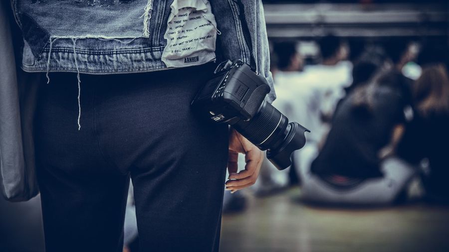 Midsection of photographer standing with camera against people sitting in room