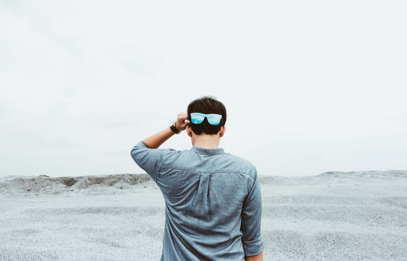 Rear view of man wearing sunglasses standing on beach against sky