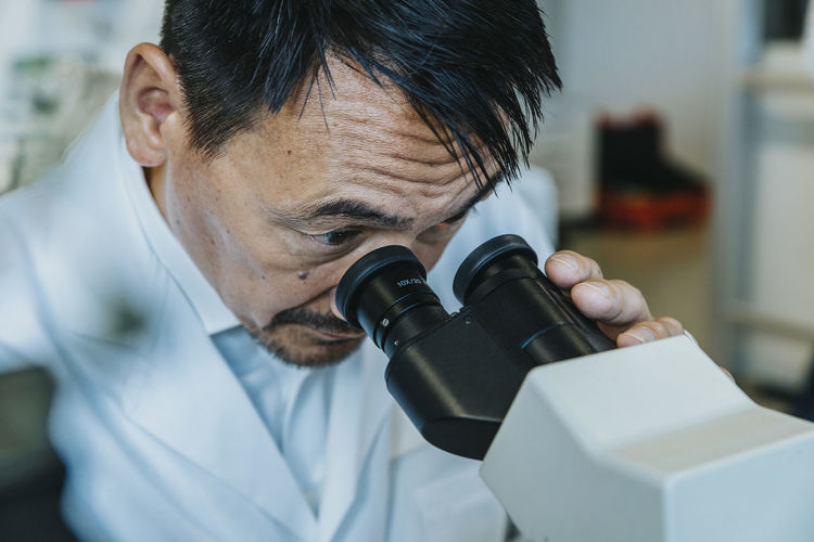Male scientist looking though microscope while man standing in background at laboratory