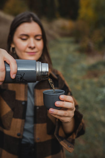 Girl wearing plaid shirt drinking tea from the thermos in forest nature