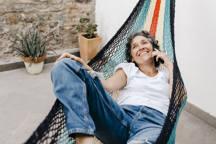 Smiling woman talking through smart phone while relaxing on hammock at back yard