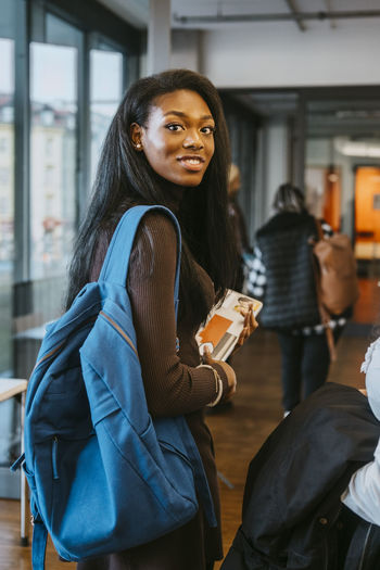 Portrait of smiling young woman holding book and backpack walking with friend at community college