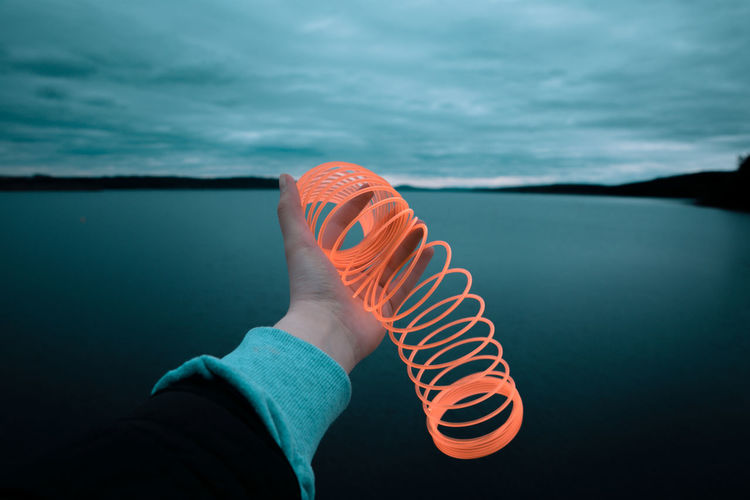 Cropped hand neon spring toy by lake against cloudy sky