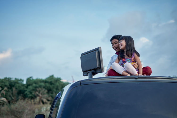 Cheerful siblings looking at desktop pc while sitting on car