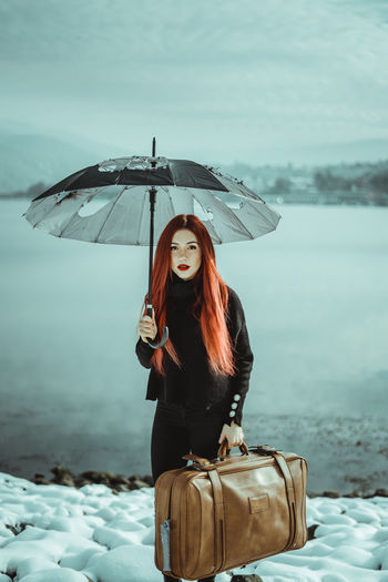 Portrait of young woman holding torn umbrella and bag while standing in snow during winter