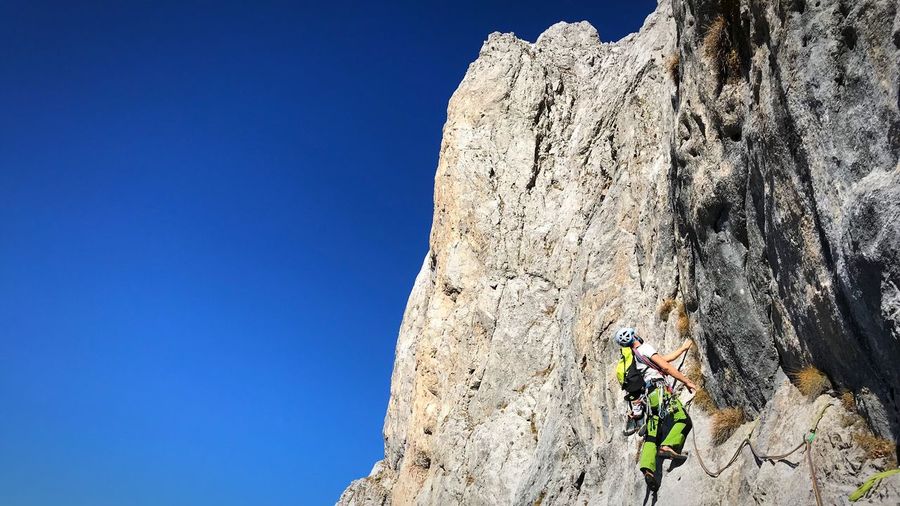Low angle view of man climbing on rock against clear blue sky