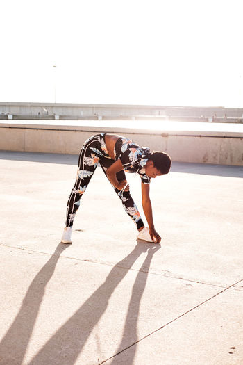 Focused african american adult woman in flowered sportswear stretching leg muscles while standing alone and warming up before training among urban environment in sunny day