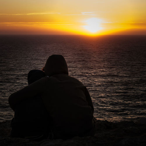 Rear view of two men sitting on sea at sunset