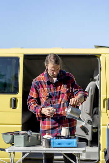 Man in checkered shirt pouring water in moka pot while preparing coffee on table against yellow camper and cloudless blue sky in morning
