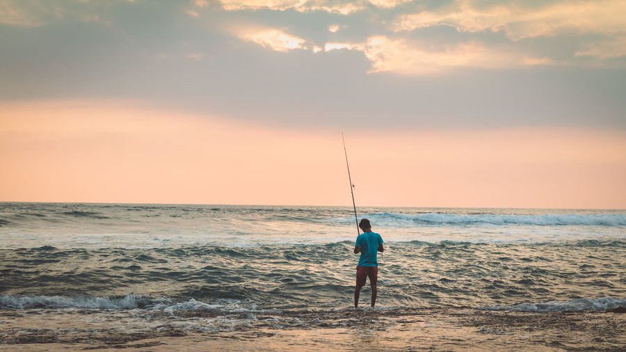 Rear view of man fishing in sea against sky during sunset