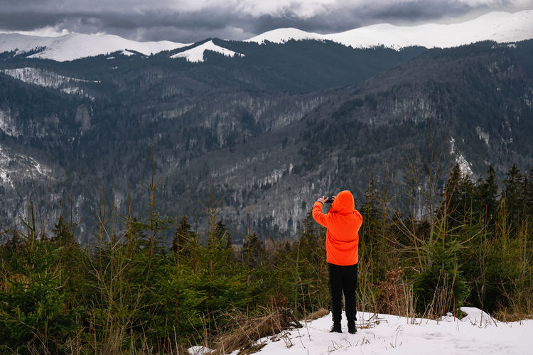 Rear view of person in orange winter jacket taking photos on snowcapped mountain during winter