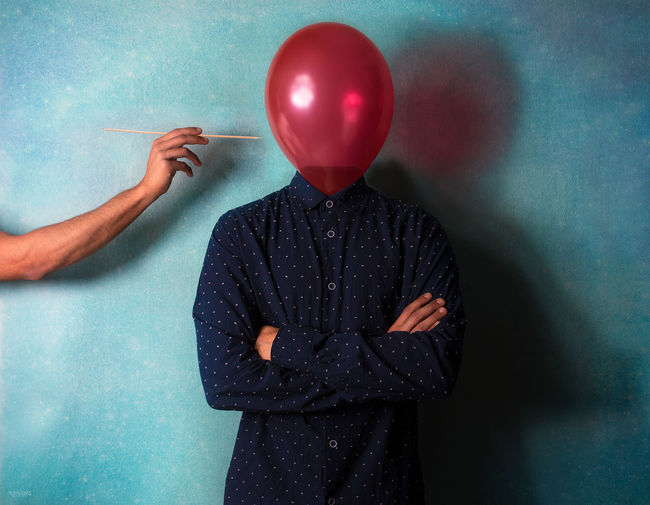 Digitally generated image of man poking balloon face with stick against turquoise wall