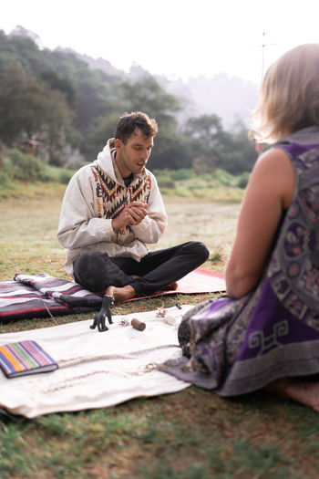 Adult female shaman with blond hair in casual clothes sitting on mat with various spiritual items and talking with man with eyes closed before meditation