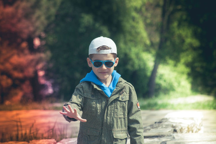 Portrait of boy with sunglasses and cap gesturing while against trees