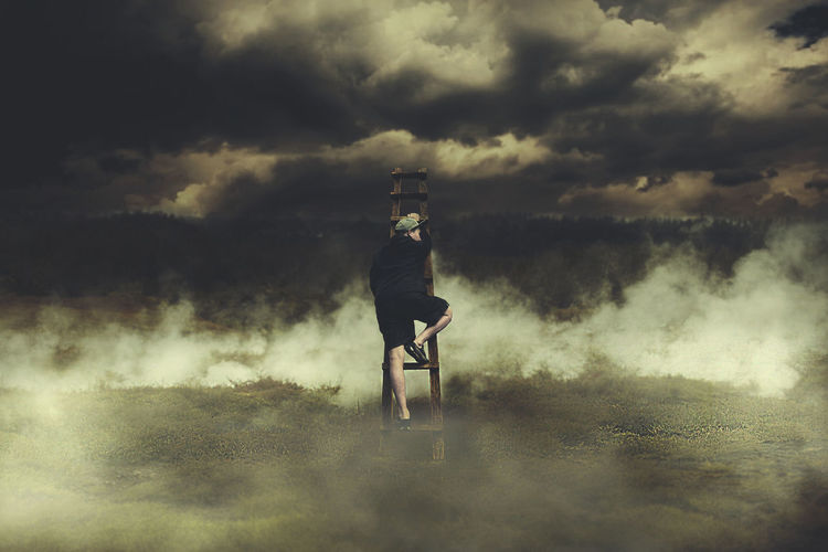 Rear view of man climbing on ladder at foggy field against cloudy sky
