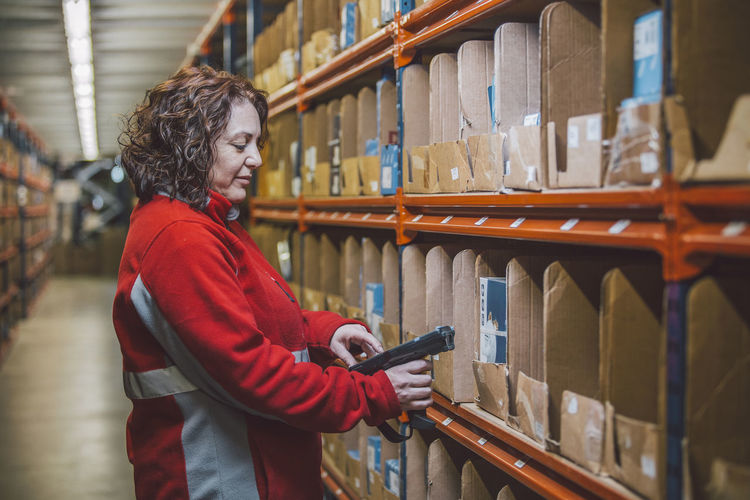 Side view of content female employee in red uniform with tool in hands standing near selves with boxes