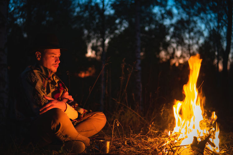 Man wearing poncho sits by the fire in the forest in dusk