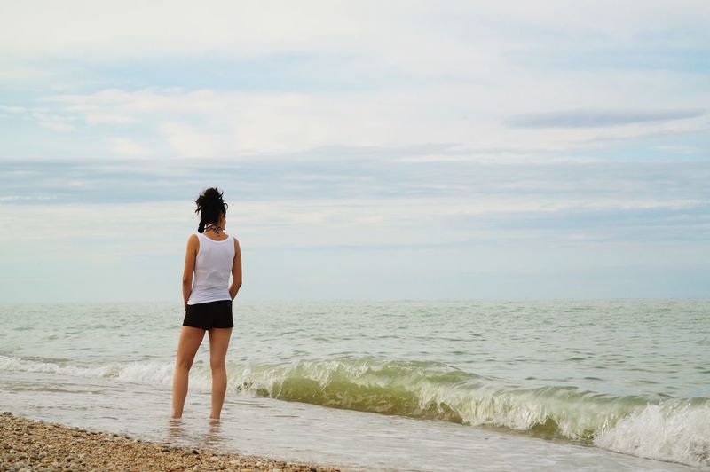 Rear view of woman standing shore at beach against cloudy sky