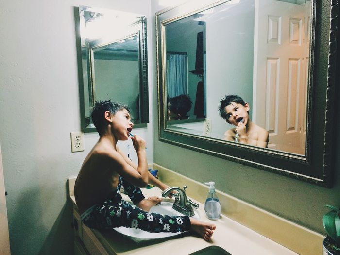 Cute shirtless boy brushing teeth in front of mirror while sitting on sink