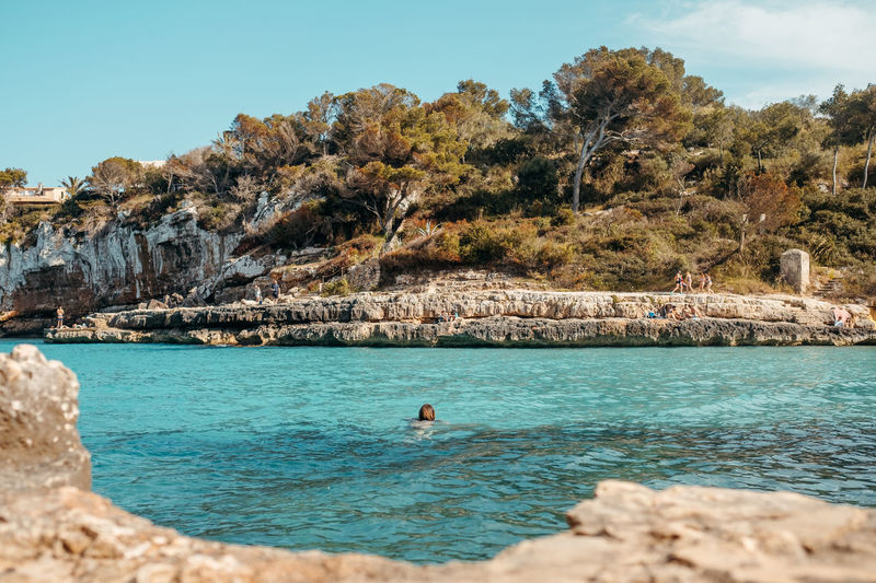 Back view of unrecognizable young female swimming on rocky seashore and admiring nature during summer holidays in mallorca