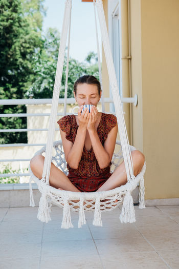 Young woman chilling in macrame chair hammock on balcony. beautiful relaxing girl drinking coffee.