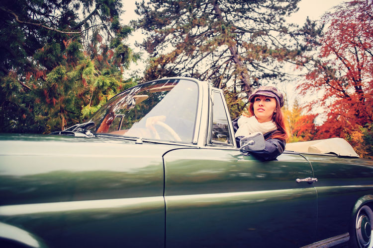 Woman wearing cap driving convertible car against trees