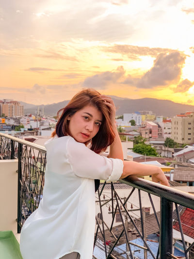 Portrait of woman standing at balcony against sky during sunset