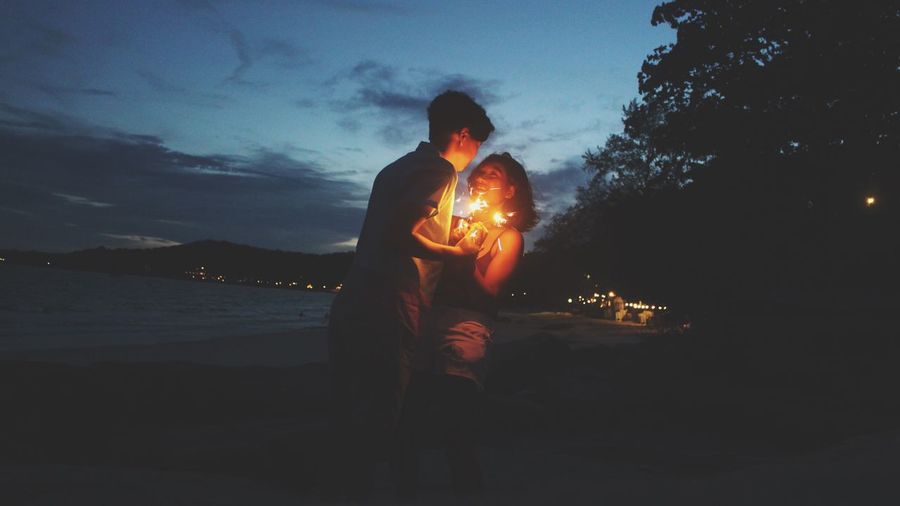 Young couple with illuminated sparklers standing at beach during dusk