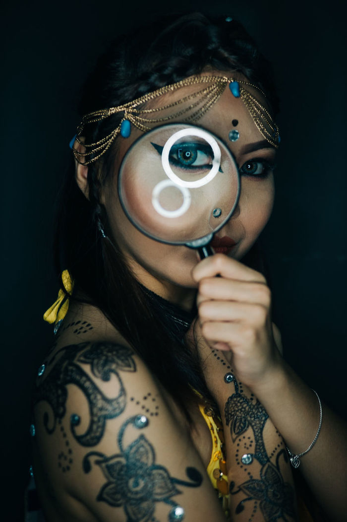 Close-up of tattooed woman looking through magnifying glass against black background