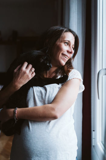Young pregnant woman at home holding a black cat on shoulder