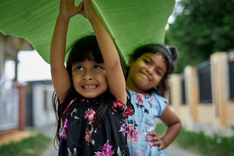 Smiling girl with arms raised holding banana leaf by sister