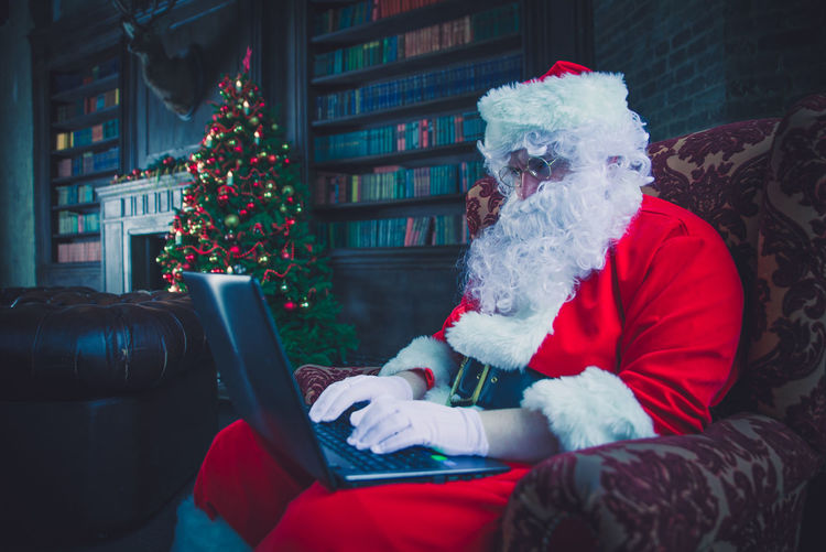 Man wearing santa claus costume using laptop on armchair at home