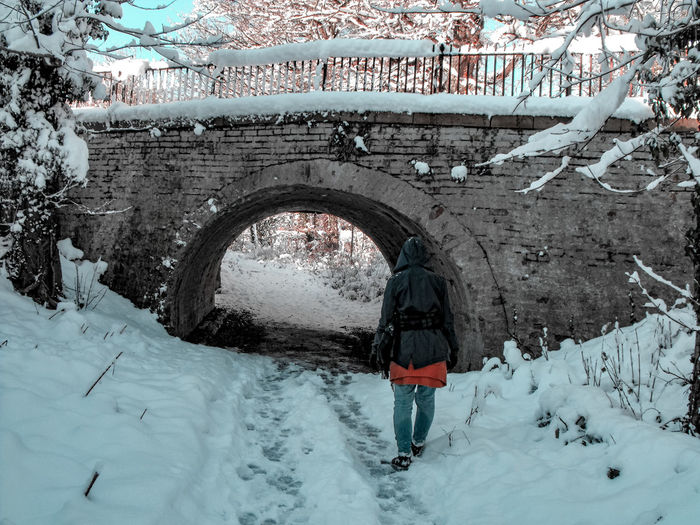Rear view of person standing in snow covered landscape by bridge