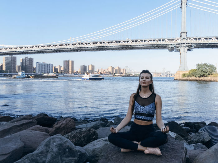 Young woman meditating while sitting on rock against bridge in city
