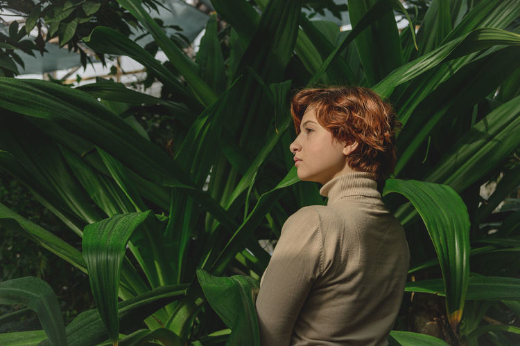 A beautiful plus size girl with red hair among the lush green branches of tropical plants. 