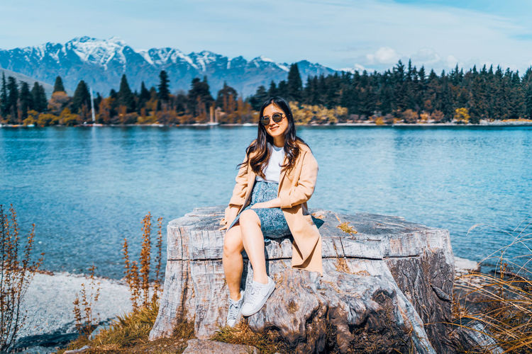 Portrait of young woman sitting on tree stump by lake against mountains and sky