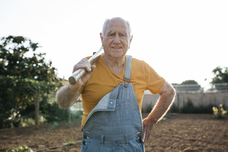 Senior man in denim overall holding garden tool on shoulder and looking at camera