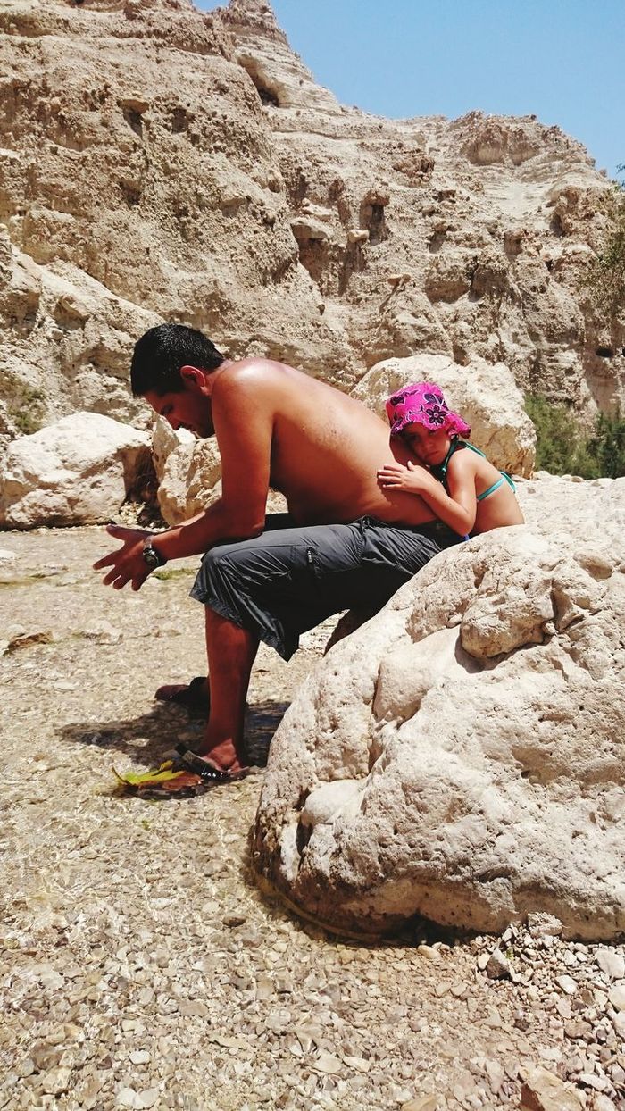 Daughter leaning over father while sitting on rock during sunny day
