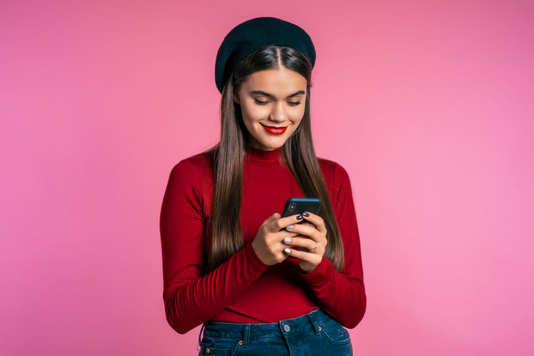 Young woman using mobile phone while standing against pink background
