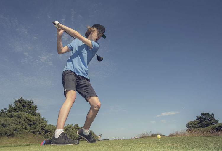 Teenage boy hitting golf shot against blue sky at course