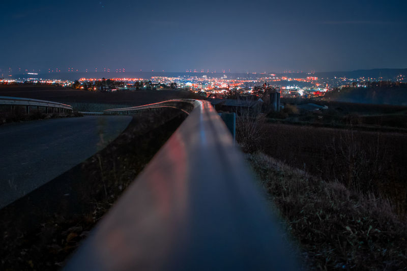 Light trails on road by illuminated city against sky at night