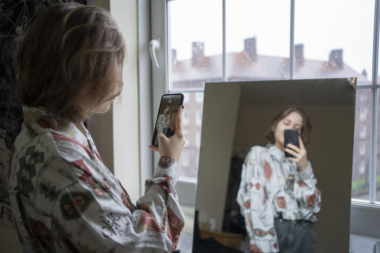 Young woman photographing reflection in mirror through smart phone at home